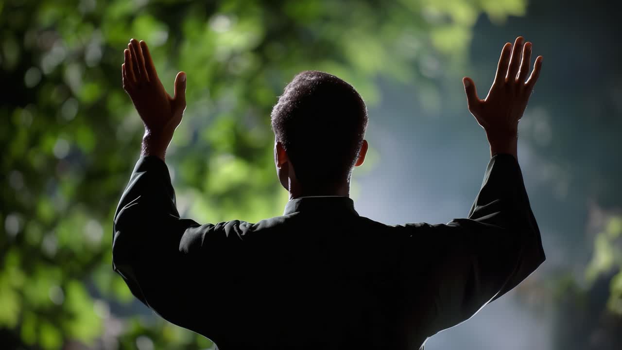 A person stands with raised hands in an impressive display of gratitude or prayer, silhouetted against a breathtaking backdrop of vibrant green foliage and soft lighting