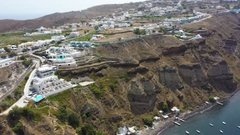 Antena-De-Lujosas-Mansiones-En-Un-Acantilado-Con-Vistas-Al-Mar-Mediterráneo-Con-Agua-Azul-Cristalina-Y-Playa-En-Santorini,-Grecia