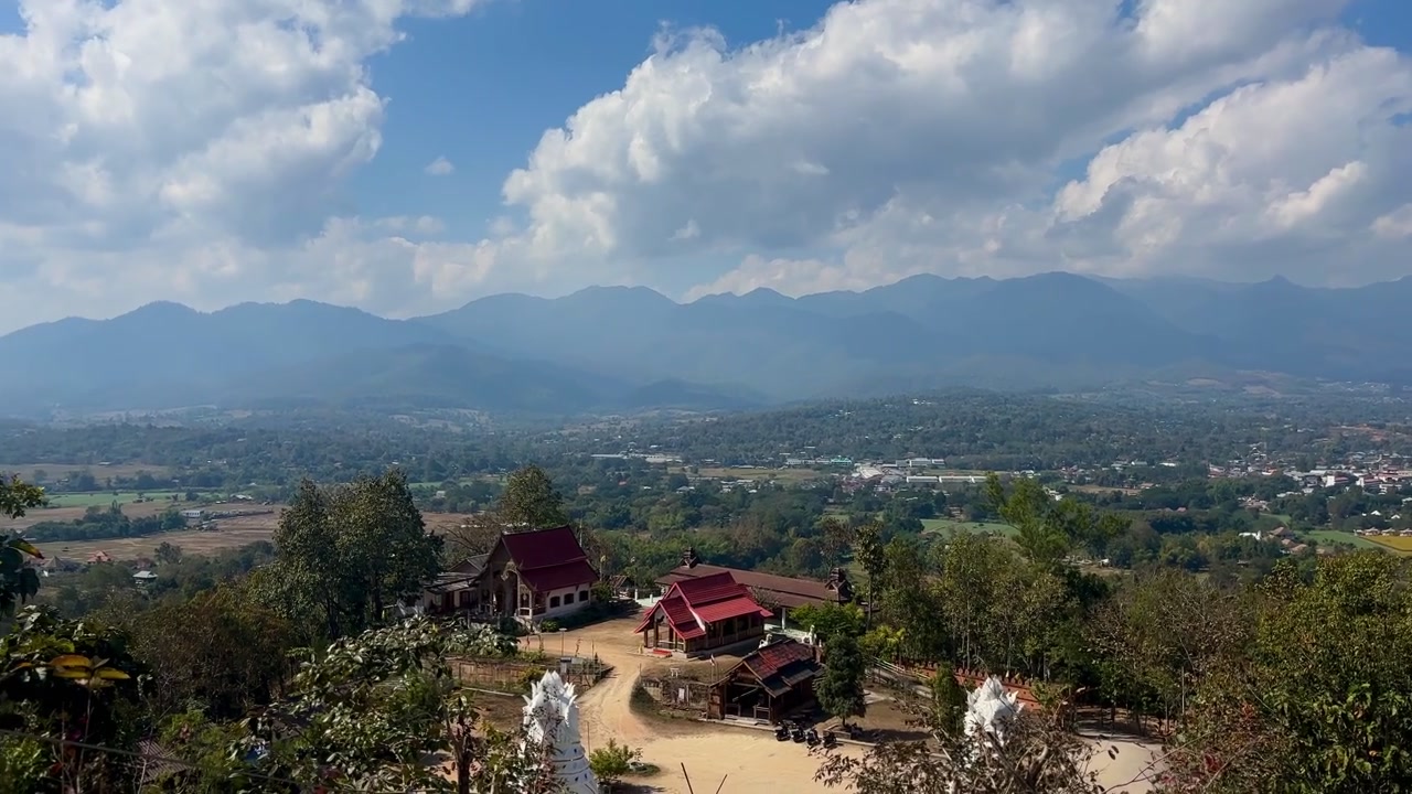 Panorering over Pai-dalen fra Wat Phra That Mae Yen i Thailand, der viser tempelbygninger med rødt tag, marker, træer og bjergkæder under dramatiske skyer