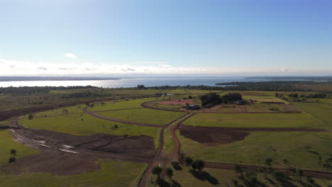 a vast green field with winding dirt roads, an equestrian field and a river in the distance, aerial view