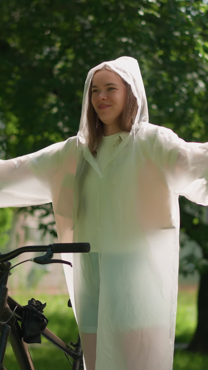 반투명한 레인코트를 입은 젊은 여성이 자전거 에 서서 팔을 기게 펴고, 마치 자유를 포옹하는 것처럼 손을 흔들고, 빛은 울창한 초록색을 통해 필터링합니다.