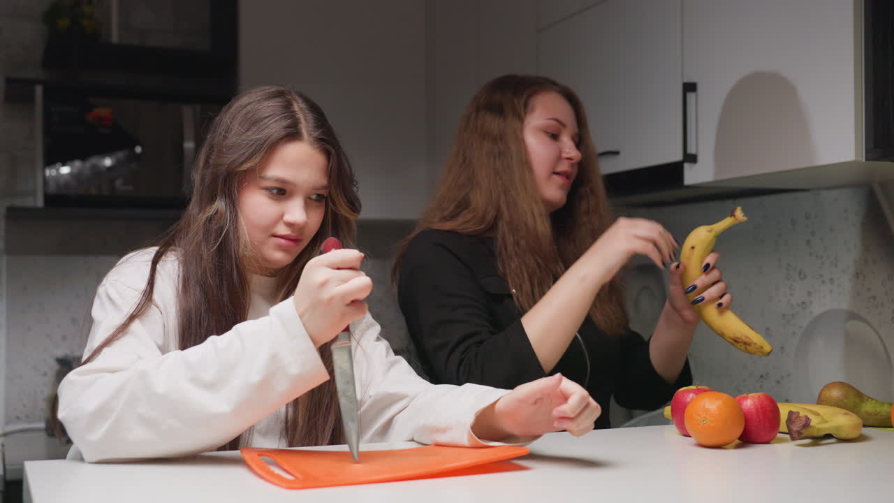 Picnic friends in kitchen, one holds chopping board with knife while other arranges fruits on table, casual cooperation, clean counter, soft light, playful preparation mood before snack time
