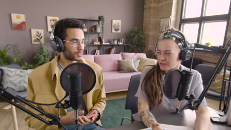 Front-View-Of-Young-Man-And-Woman-Wearing-Headphones-Sitting-At-A-Table-With-Microphones-While-They-Recording-A-Podcast