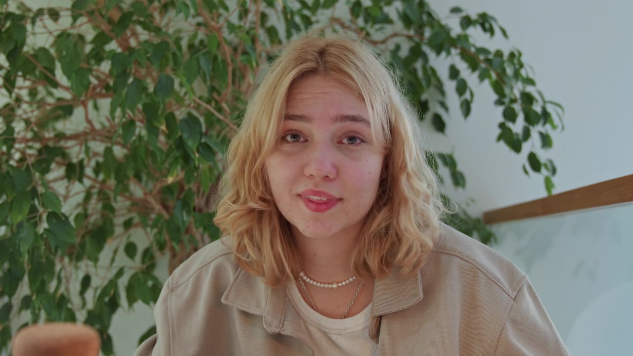 Close up of young woman with blonde hair wearing layered necklaces and open jacket seated in front of green foliage. scene is calm indoors with soft lighting featuring natural textures