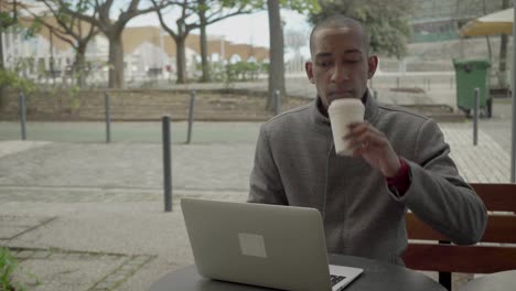 Un-Joven-Con-Abrigo-Usando-Una-Computadora-Portátil-Y-Bebiendo-De-Un-Vaso-De-Papel