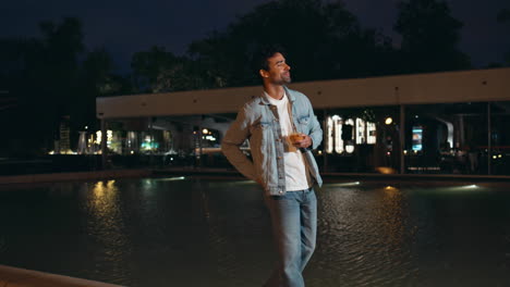 carefree guy walking night area near water pool. handsome man holding glass