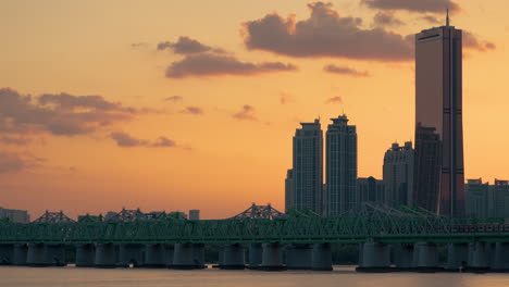 Seoul-Subway-Passenger-Train-Moves-on-Green-Railway-Bridge-over-Hangang-During-Sunset-with-Dramatic-Orange-Sky,-63-Building-Skyscraper-Towering-in-Backdrop,-copy-space-for-text