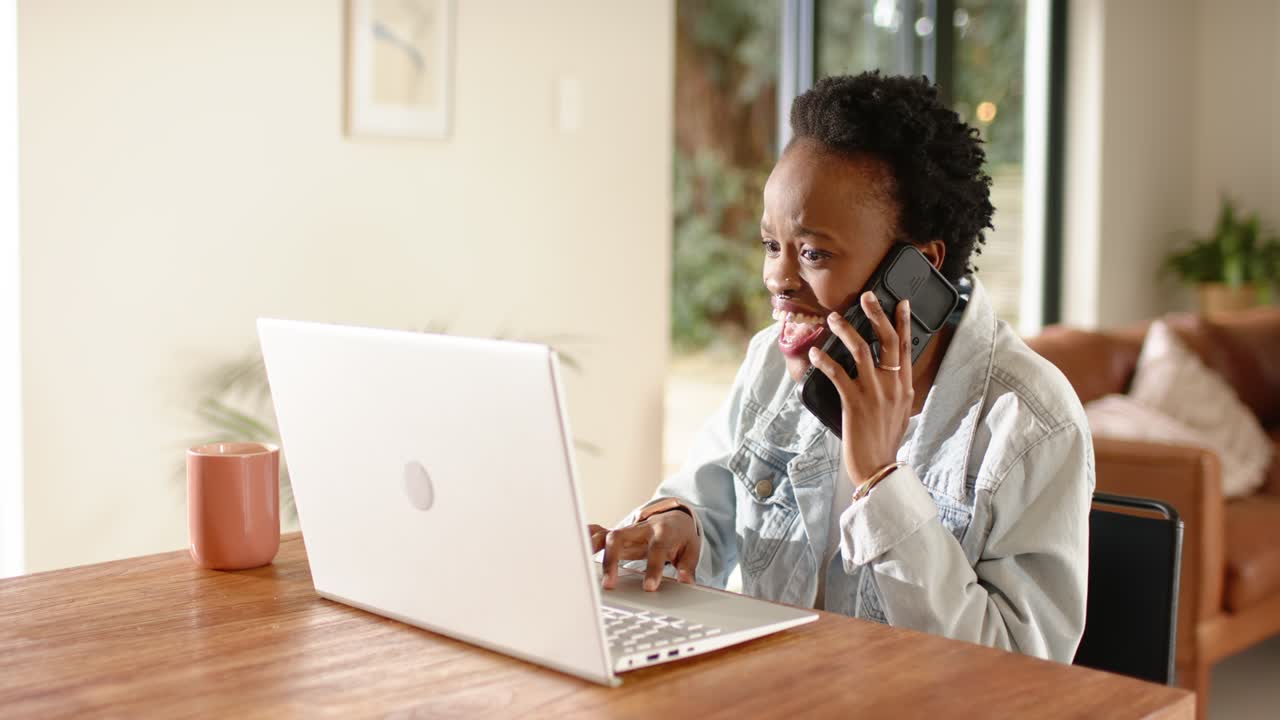 Afro-Amerikaanse vrouw beantwoordt smartphone, voert gegevens in op laptop in thuiskantoor met koffiemok.