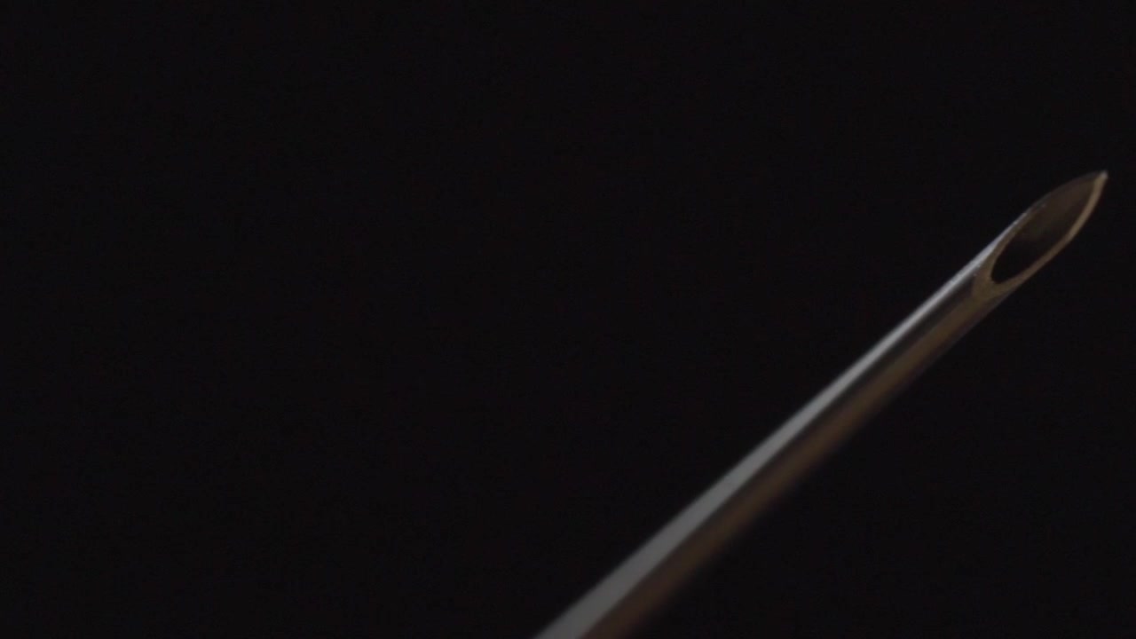 Macro primo piano della punta di un ago ipodermico su sfondo scuro. L'apertura smussata cava e la superficie metallica lucida rivelano dettagli precisi dello strumento medico.