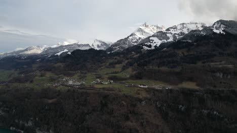 Panorámica-Aérea-A-Través-De-Casas-Que-Salpican-La-Ladera-Cubierta-De-Hierba-En-La-Meseta-Sobre-El-Lago-Walen-Con-Picos-De-Montañas-Cubiertas-De-Nieve