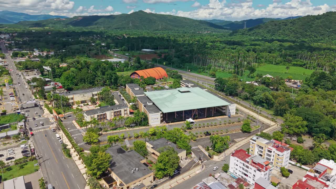 Aerial orbit of Autonomous University in Bonao with green mountains in background. Sunny day with clouds in Dominican Republic. Modern university infrastructure with pyramidal roof