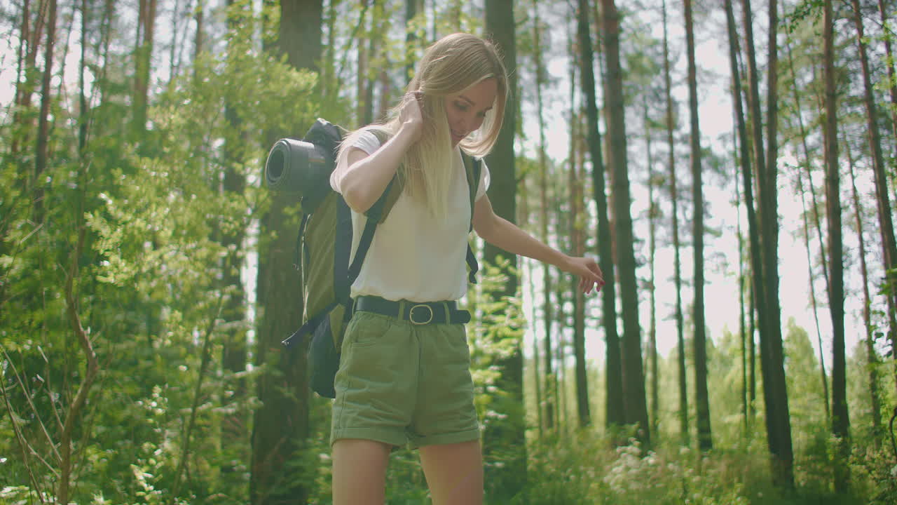 젊은 여성 금발 관광객이 모험 중에 로그를 통과하는 배 ⁇ 과 함께 느린 움직임으로 숲을  ⁇ 고 있습니다.