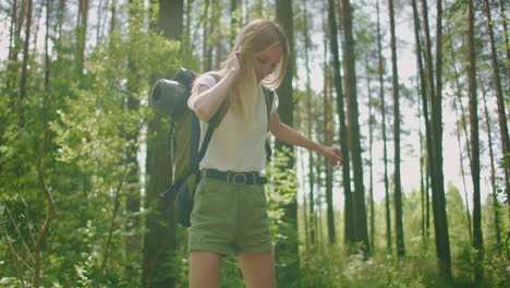 a young female blonde tourist walks through the woods in slow motion with a backpack passing through a log during an adventure