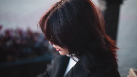 close-up of woman with soft, flowing brown hair and thoughtful expression in outdoor urban environment, background features blurred plants and subtle light tones