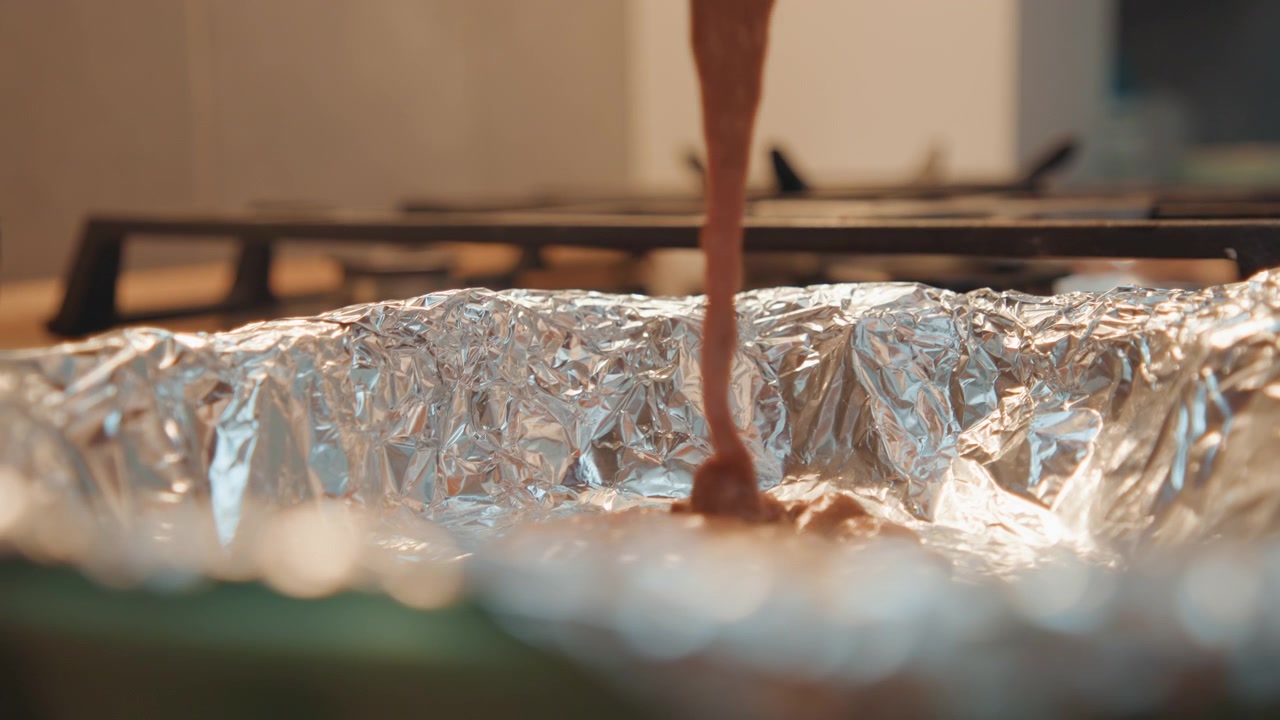 Un chorro oscuro y brillante fluye hacia una fuente para hornear. Una rica mezcla de chocolate llena suavemente y con elegancia una bandeja para hornear preparada. Se vierte una masa de chocolate sedosa en el molde, creando una capa brillante y deliciosa.