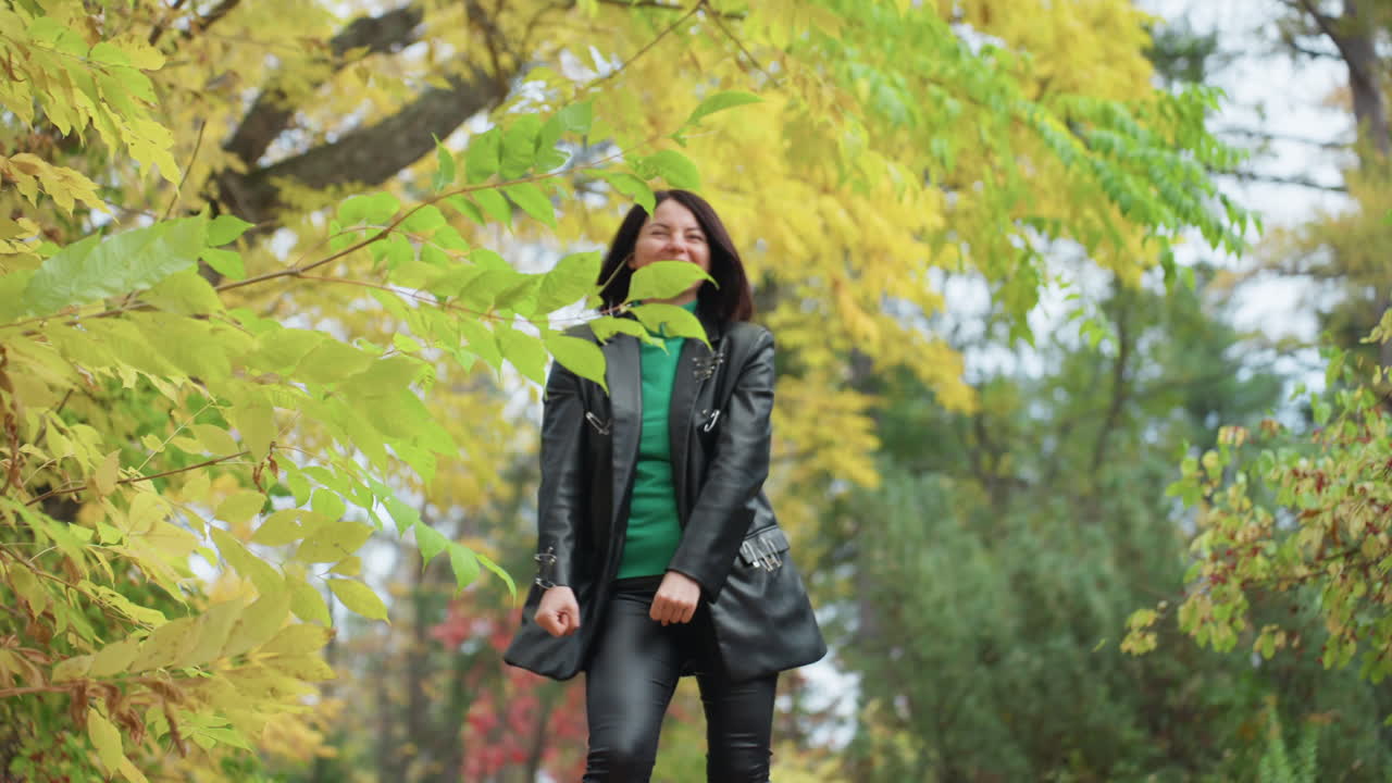 woman in leather jacket walking through yellow autumn trees wearing green turtleneck, smiling and tossing leaves in park candid fashion portrait with rock edge and relaxed confident mood, soft