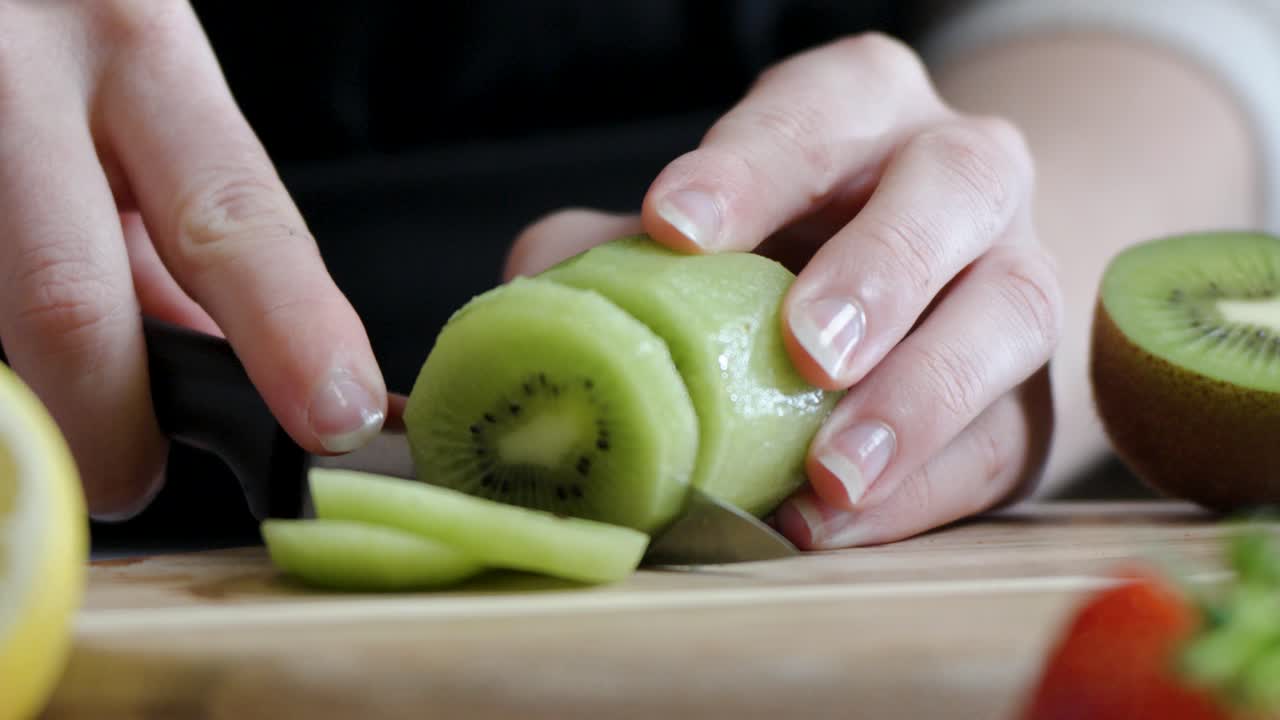Hand cuts peeled kiwi into thin slices surrounded by other fresh fruits