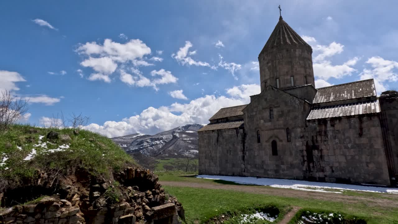 Visitors walk around an ancient monastery in Armenia while enjoying the clear skies and snow on the ground. The mountains provide a stunning backdrop to this historical site