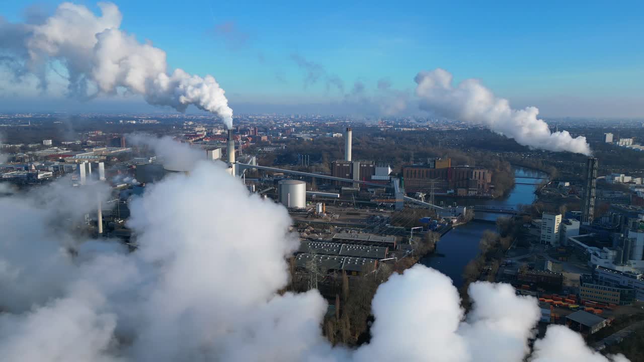 Aerial view of a thermal power plant emitting smoke on a sunny day, with a river and a city in the background. Perfect aerial view flight overflight flyover drone