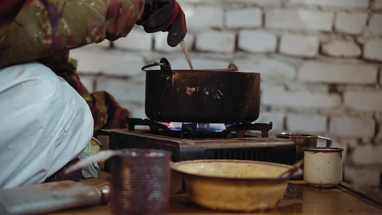 Close up of individual preparing soup in old pot on gas stove inside abandoned shelter, stirring meal with spoon, surrounded by worn cups and dishes, symbolizing survival