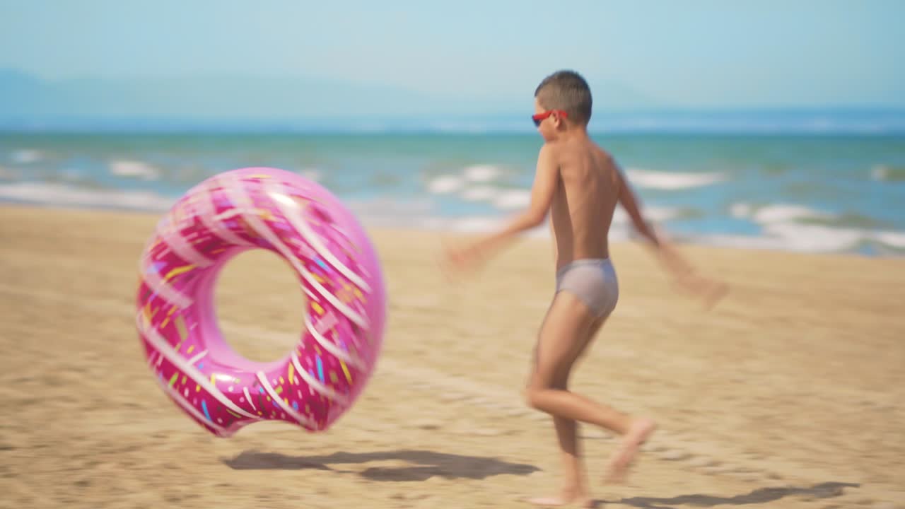 el niño corre a lo largo de la playa con una rosquilla inflable rosada, la enrolla a lo longo de la arena contra el fondo del mar. el concepto de relajación y diversión