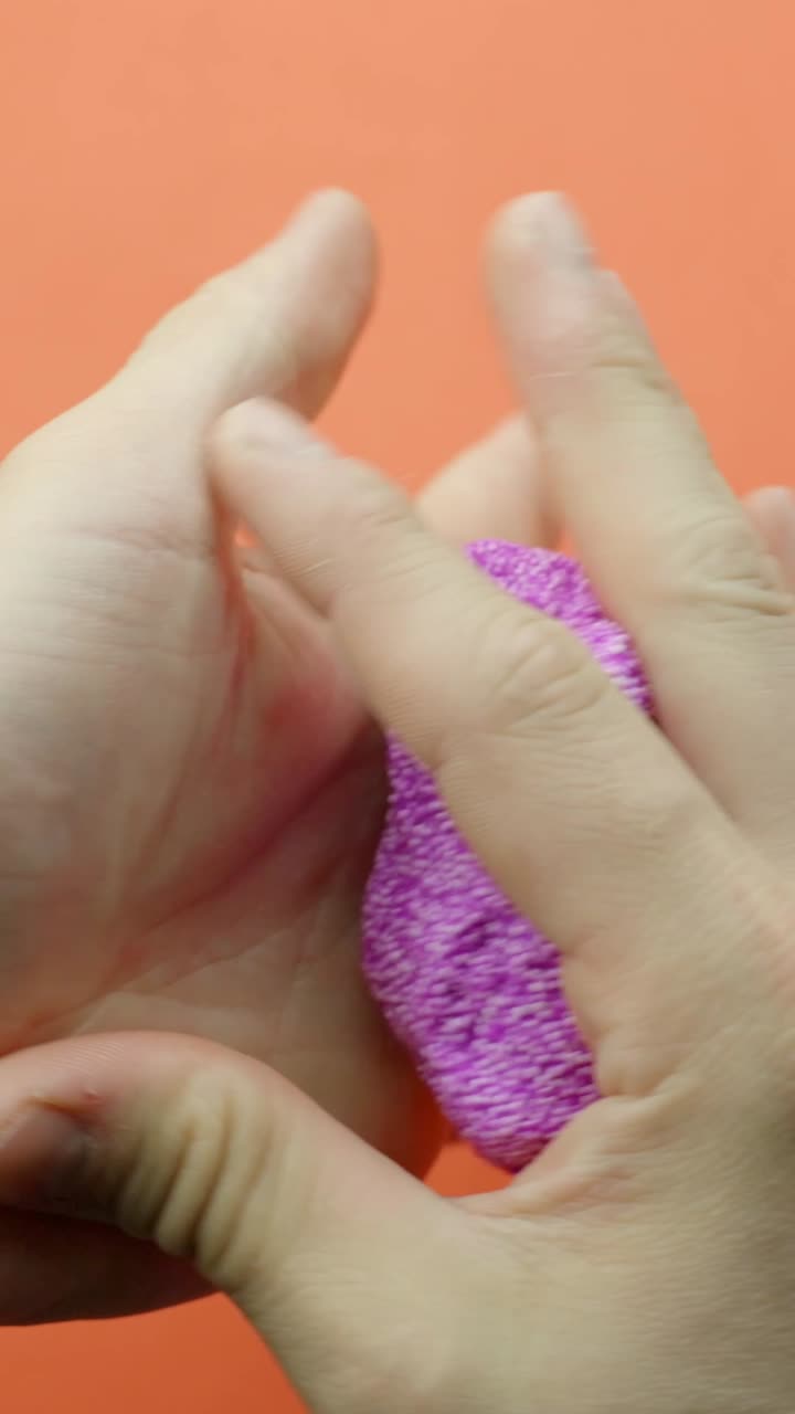 Close-up of hands pulling and stretching textured purple foam slime against an orange background. Vertical