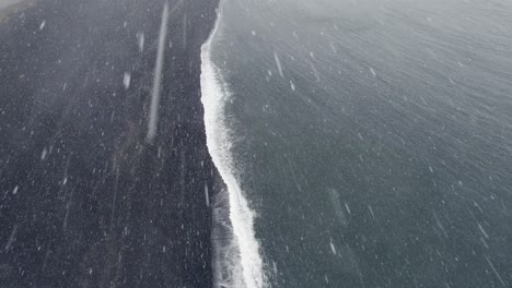 Heavy-snow-falling-over-black-sand-beach-in-Iceland,-High-angle-drone-view