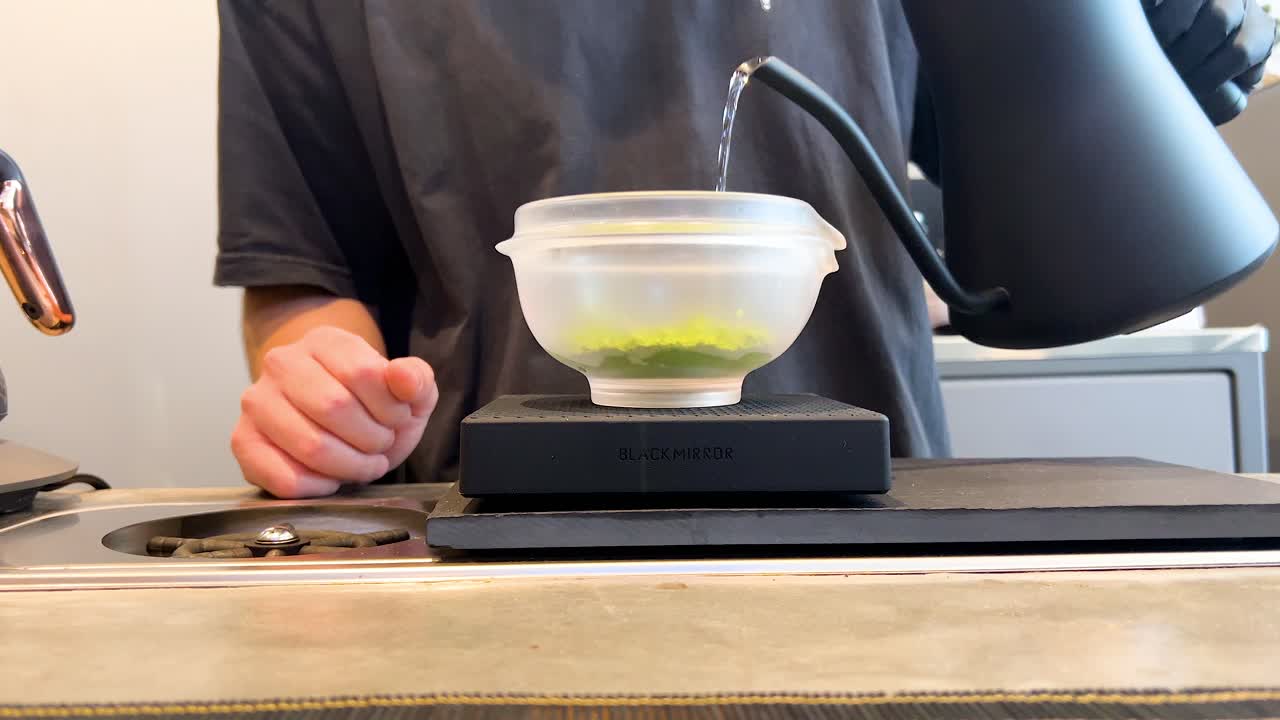 Barista pours hot water into matcha bowl, bright indoor lighting, steady camera, contemporary café setting