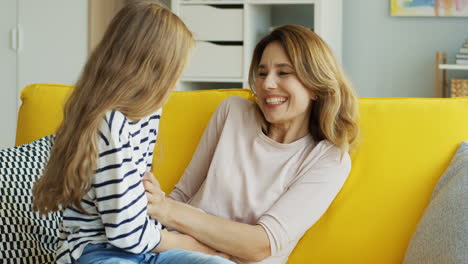 Hermosa-Madre-E-Hija-Adolescente-Divirtiéndose-En-Un-Sofá-Amarillo-Y-Haciéndose-Cosquillas-En-La-Sala-De-Estar