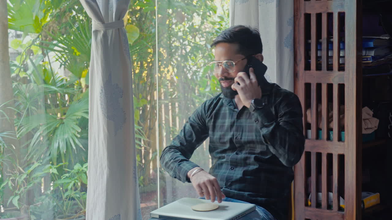 Indian young man working on laptop and talking on mobile phone near window side at home.