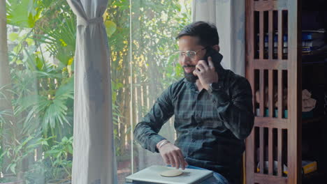 indian young man working on laptop and talking on mobile phone near window side at home.