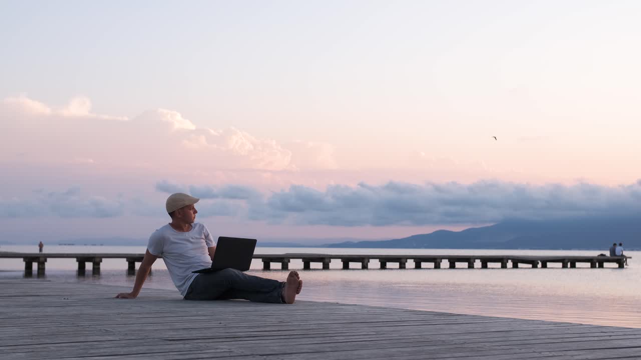 Male traveler sitting on the shore using laptop. Digital nomad or remote worker concept