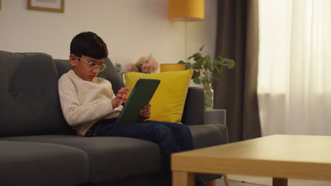 young boy sitting on sofa at home playing games or streaming onto digital tablet 9