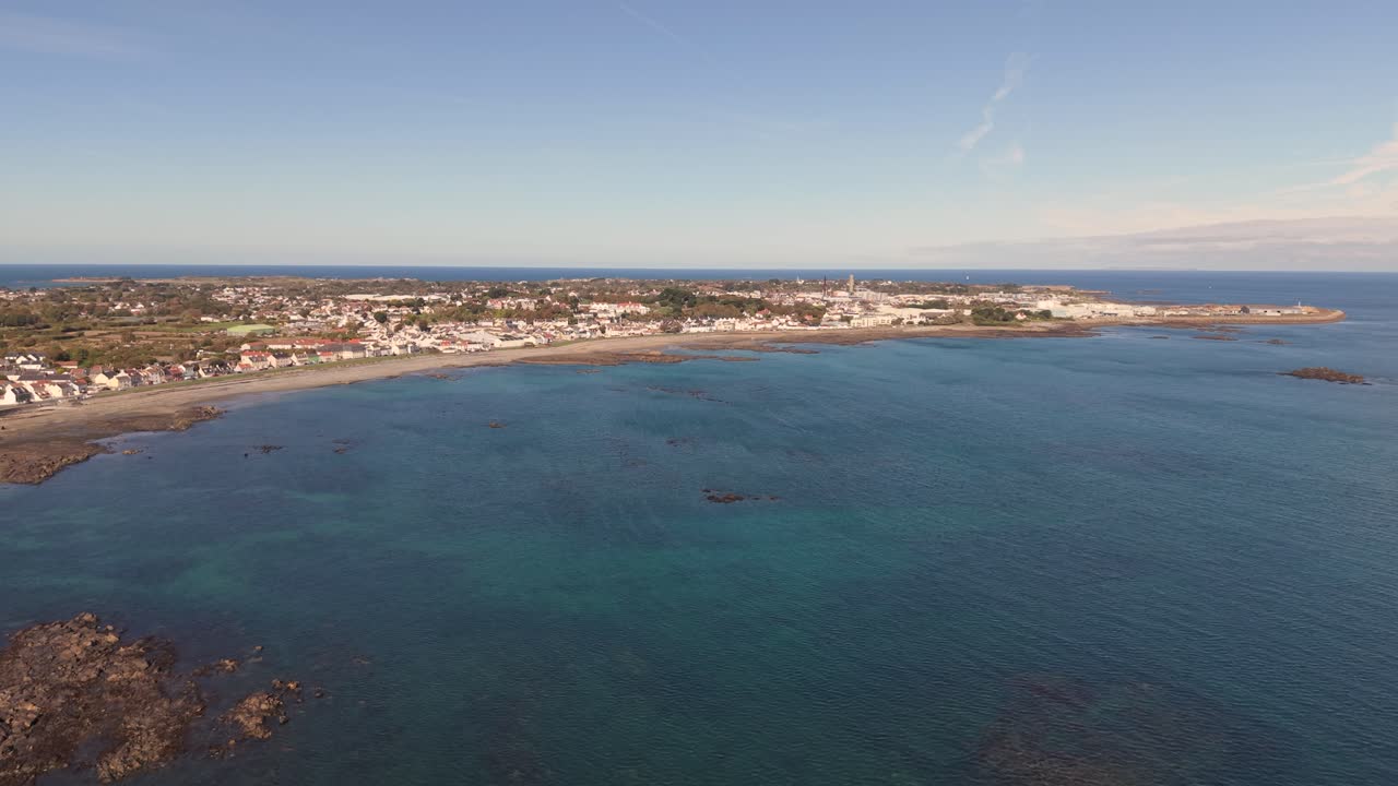 High slowly dropping drone footage over Belle Greve Bay Guernsey at high tide showing beach,shoreline,rocks,clear blue water and views of St Sampson on sunny day