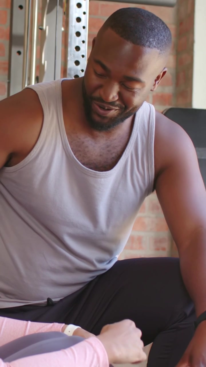 Vertical video: Leaning trainer wearing grey tank guiding trainee reaching up on bench, weight rack