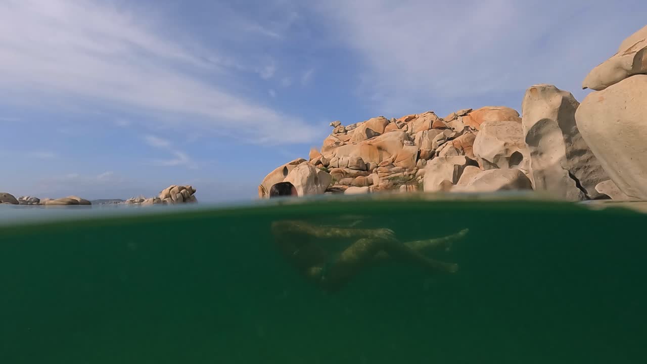 Halvt undervandsoptagelse af en kvinde, der slapper af mens hun svømmer i havvandet i Cala Della Chiesa lagunen, med eroderede granitklipper i baggrunden af Lavezzi-øen på Korsika, Frankrig.