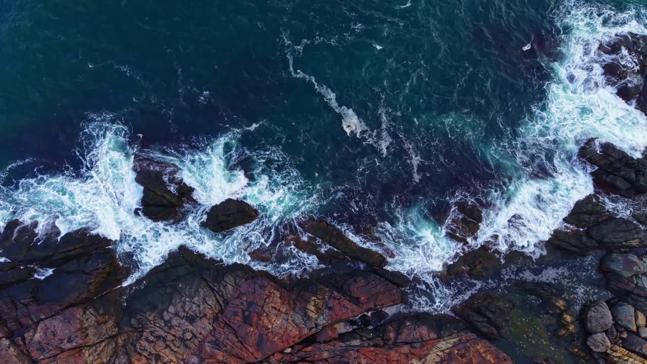 Le onde si infrangono sulle rocce frastagliate della costa oceanica sotto un cielo azzurro e un sole pomeridiano splendente.