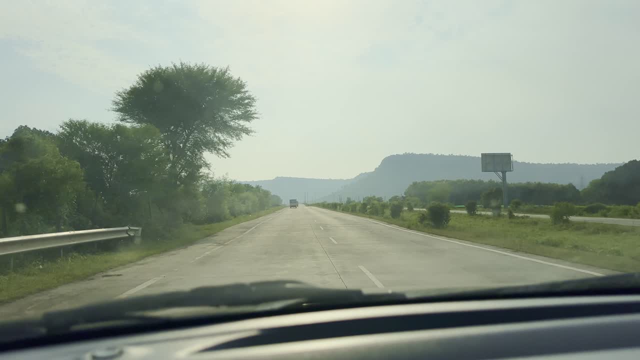 Point of view shot from inside a car traveling on a smooth Indian highway, with trees, greenery, and distant hills under bright daylight