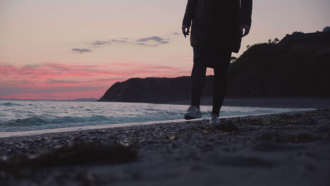 tiro de piernas en cámara lenta de una chica caminando por la playa al atardecer, con olas de mar aplastando en la orilla en el fondo