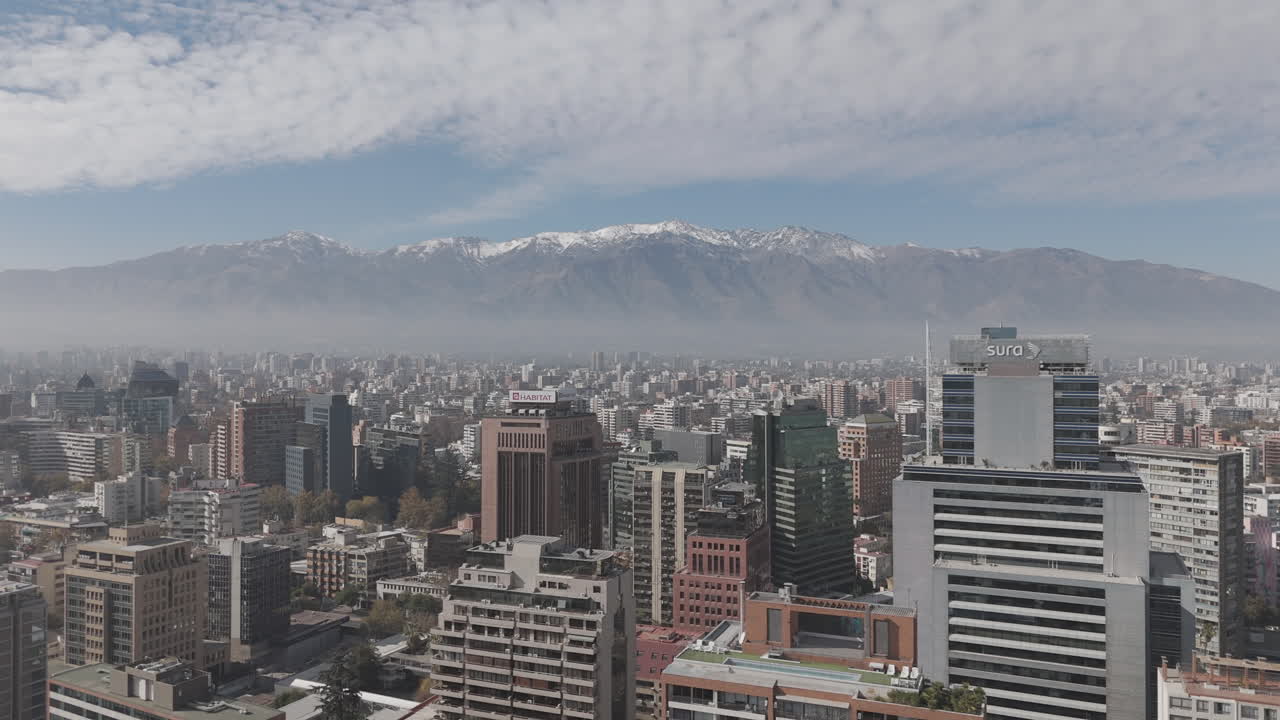 Forwards drone shot of Santiago de Chile in South America watching over the skyscrapers and big buildings with snowy mountains in the background on bright but cloudy day LOG