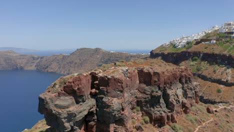 Toma-Panorámica-Aérea-De-Un-Hombre-Parado-En-La-Roca-Skaros-En-Santorini,-Grecia