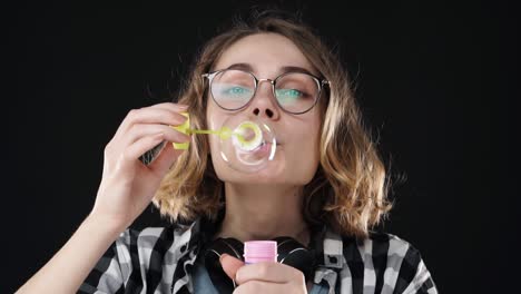 closeup portrait of happy emotional girl blowing a big soap bubble with stick on black background. studio footage of brunette with short hair and sensual lips wearing headphones on neck