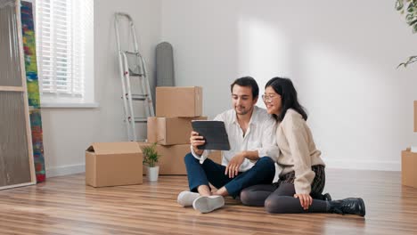 los amantes están sentados en el suelo en un apartamento recién comprado un hombre elegantemente vestido está sosteniendo una tableta una pareja está charlando con sus padres amigos por internet saludando a la cámara jactándose de mudarse a un apartamento
