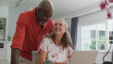 Happy-senior-diverse-couple-wearing-shirts-and-using-laptop-in-living-room