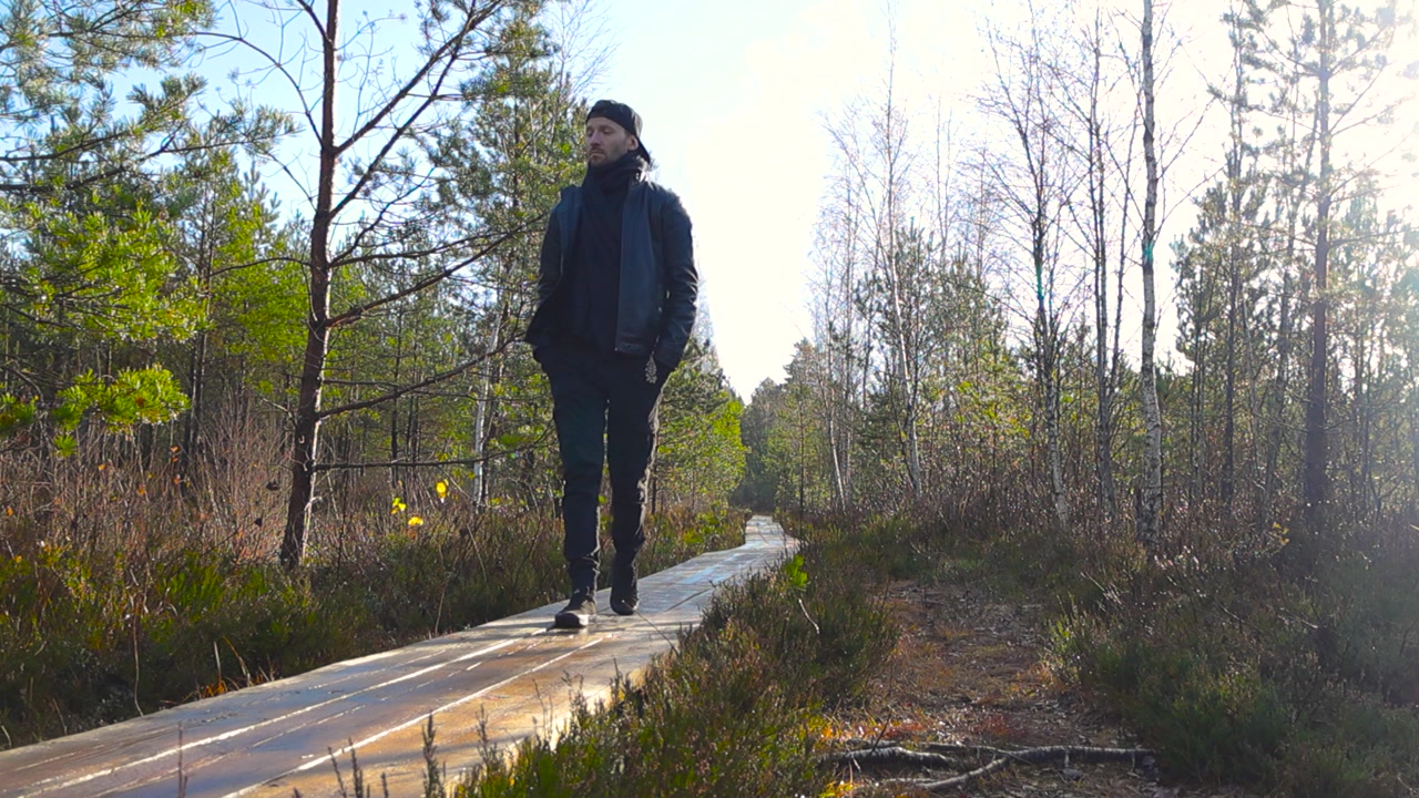 Hombre vestido de negro caminando desde la distancia hacia y más allá de la cámara en un paisaje de pantano, Estonia. El hombre lleva las manos en los bolsillos y mira a su alrededor mientras camina por el paseo de madera.
