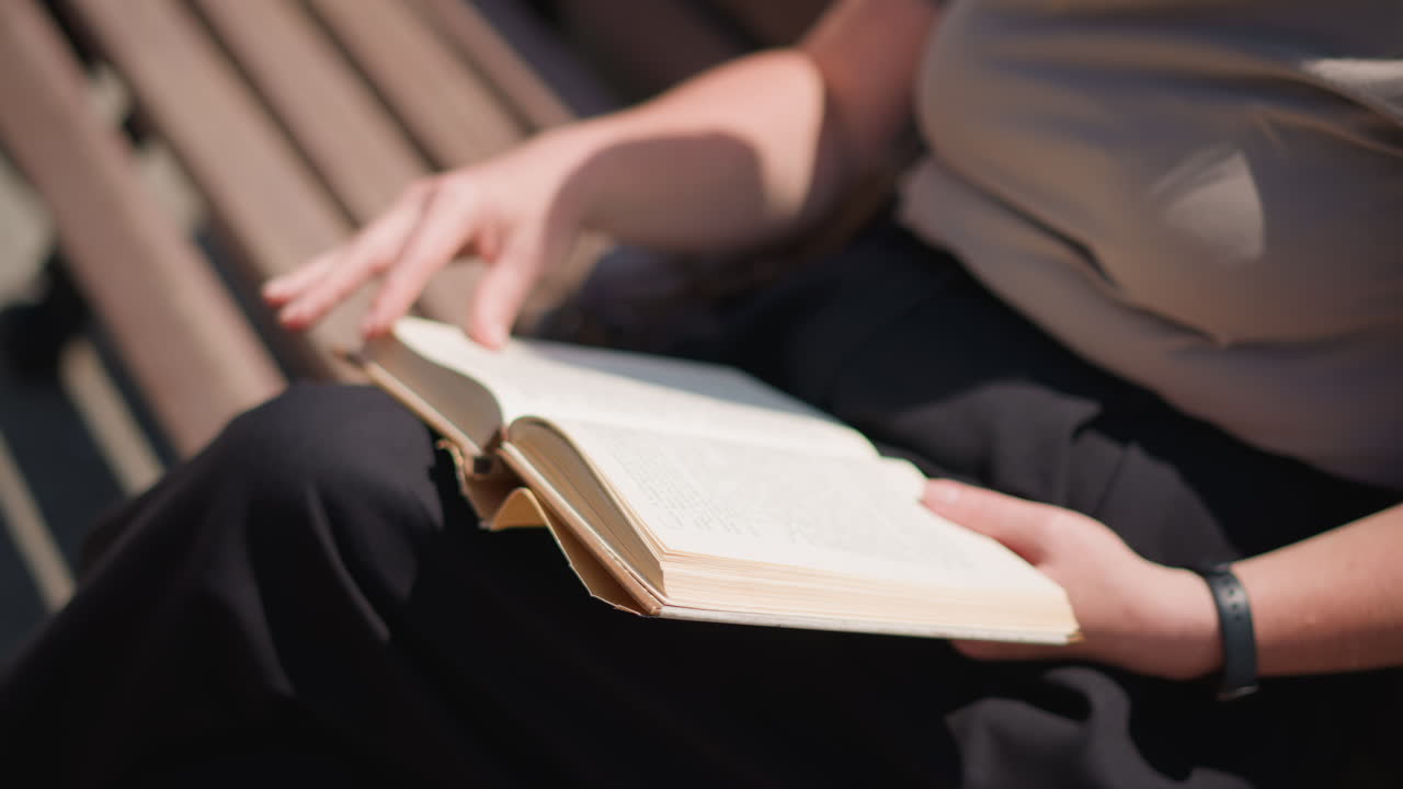 Close up of woman sitting outdoors on wooden bench flipping to new page in open book under sunlight peaceful summer mood showing calm concentration leisure study