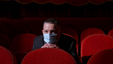 spectator in medical mask sitting alone in theater. one man in protective mask watching a play in empty auditorium during coronavirus.