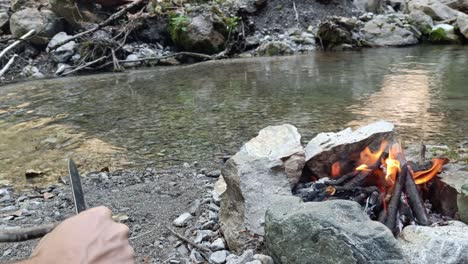 Guy-sharpen-wooden-stick-with-knife-next-to-bonfire-to-put-sausage-on-it