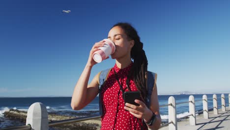 african american woman drinking coffee and using smartphone on promenade by the sea