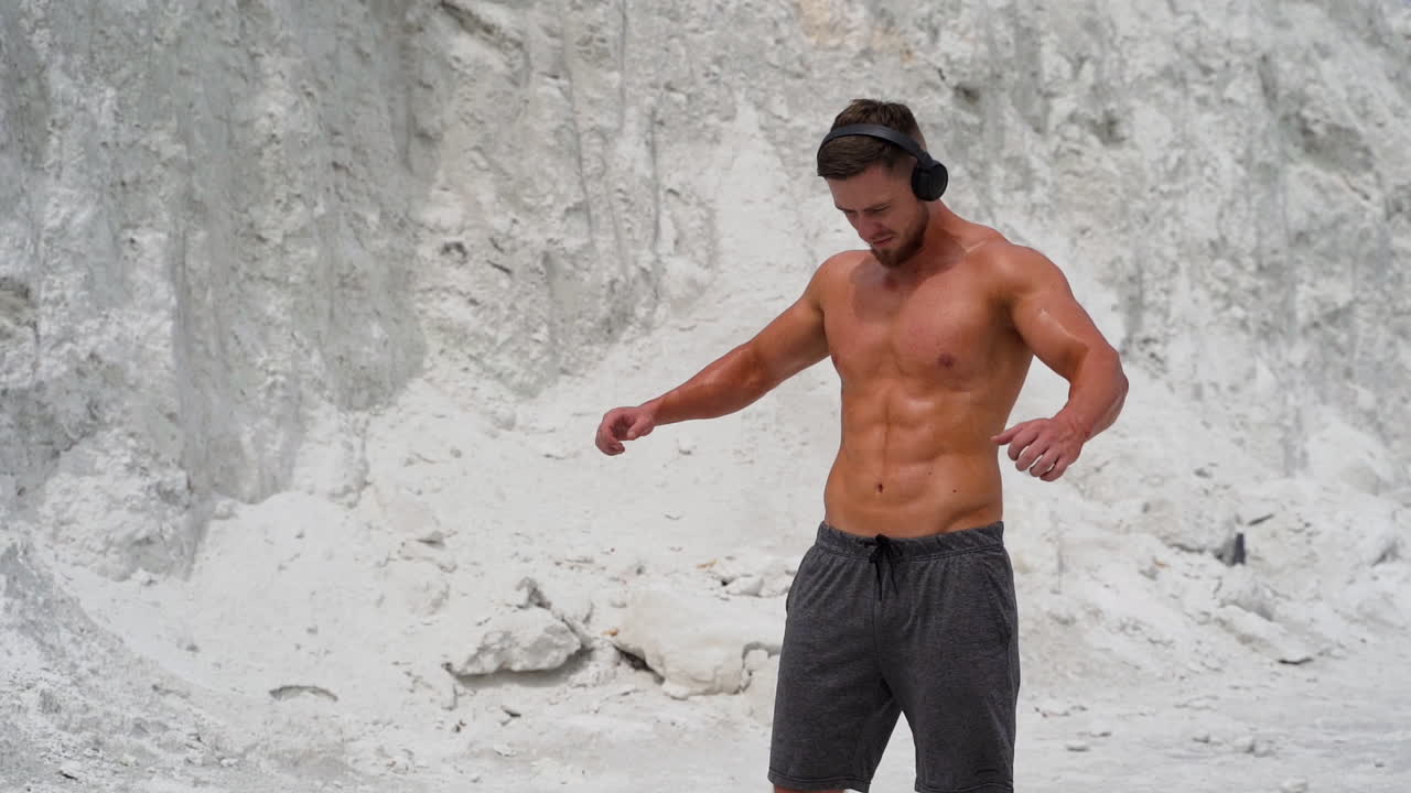 Strong man with headphones is doing exercises on the mountain background. Young shirtless male with muscular body is training in the open air in white mountains.